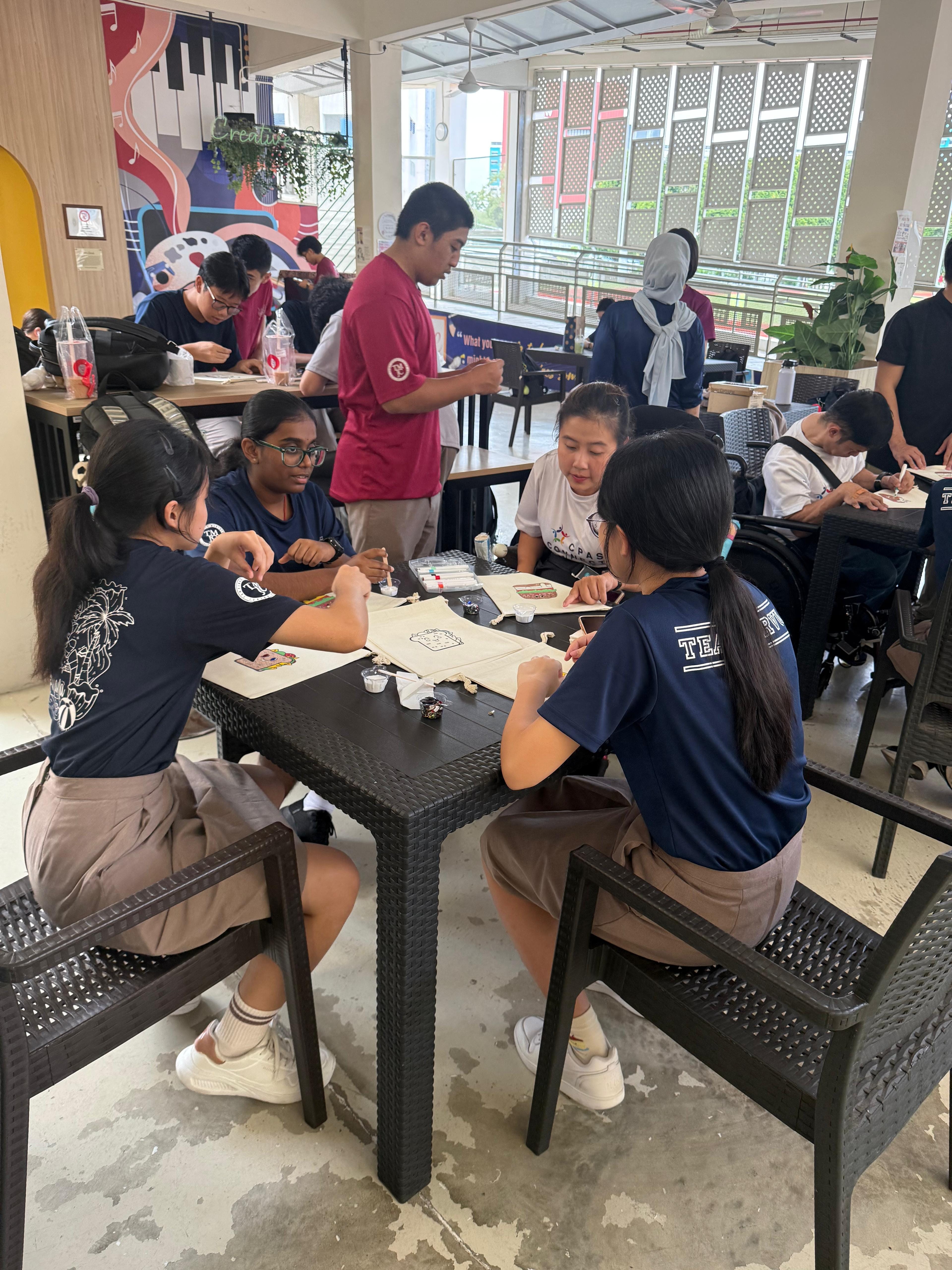 Students and CPAS alumni seated together at outdoor tables, decorating fabric items with markers and craft supplies.